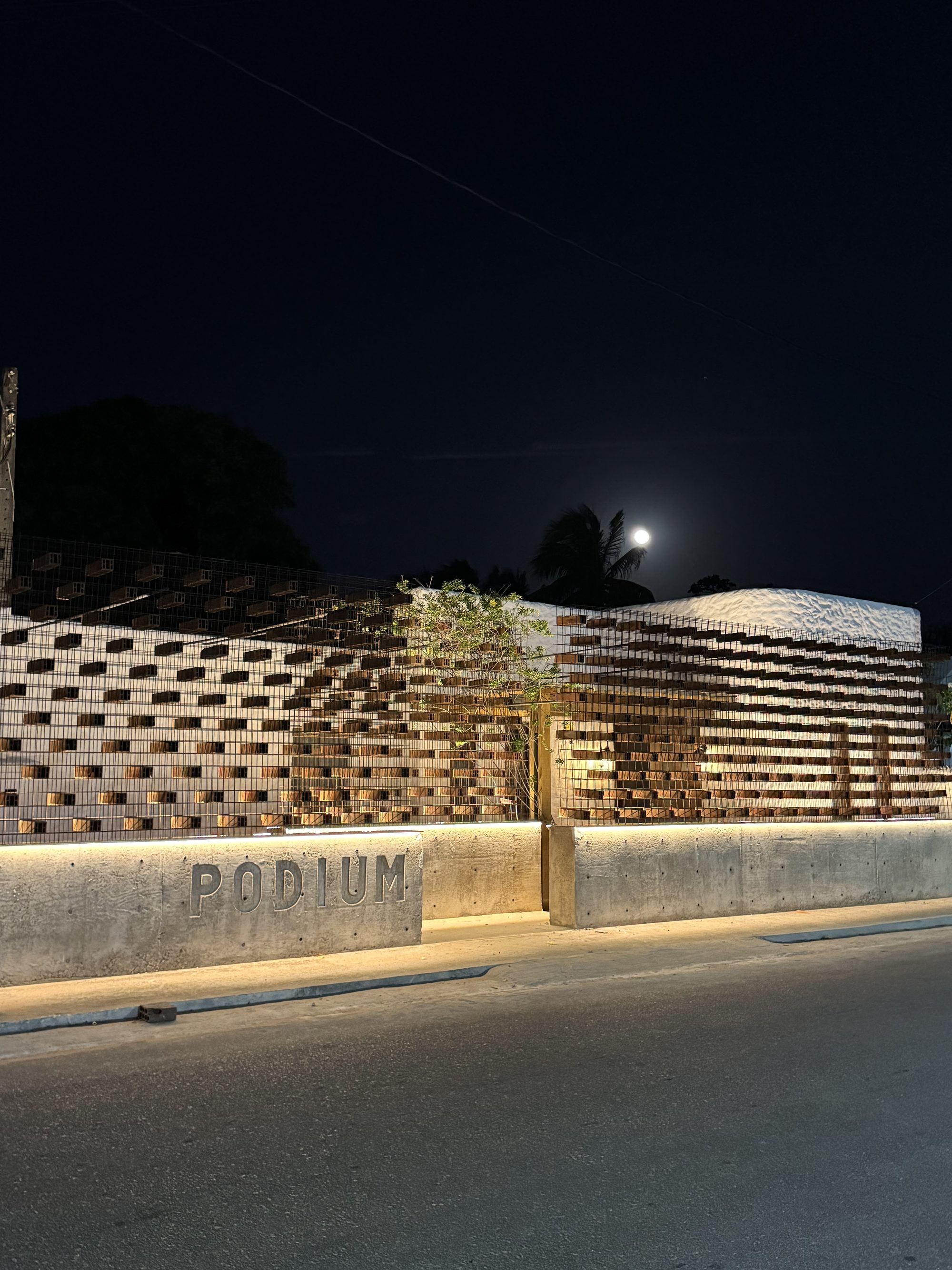 Fachada da sede da Podium em Tibau do Sul — muro de telhas cerâmicas e concreto aparente, retroiluminado à noite sob a lua